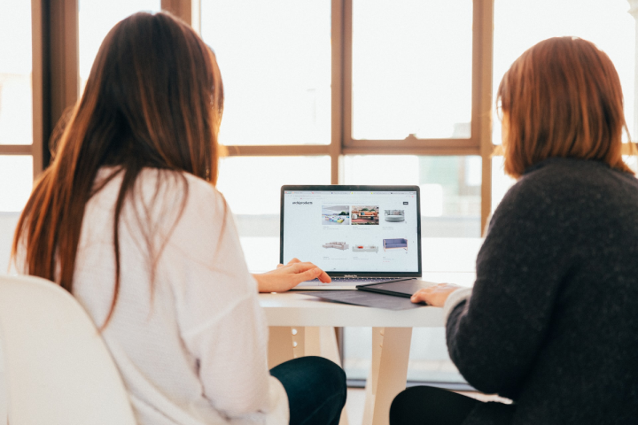 two women sitting at a table looking at a website on a laptop