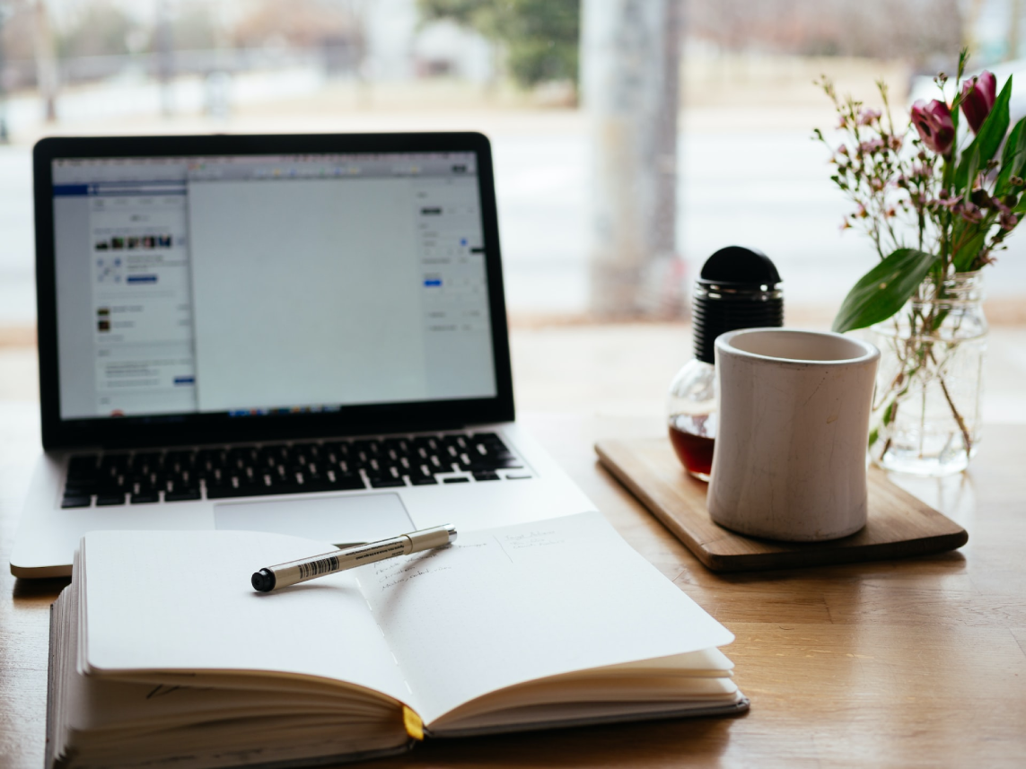 laptop, book with pen and cup of coffee with vase of flowers all on a table